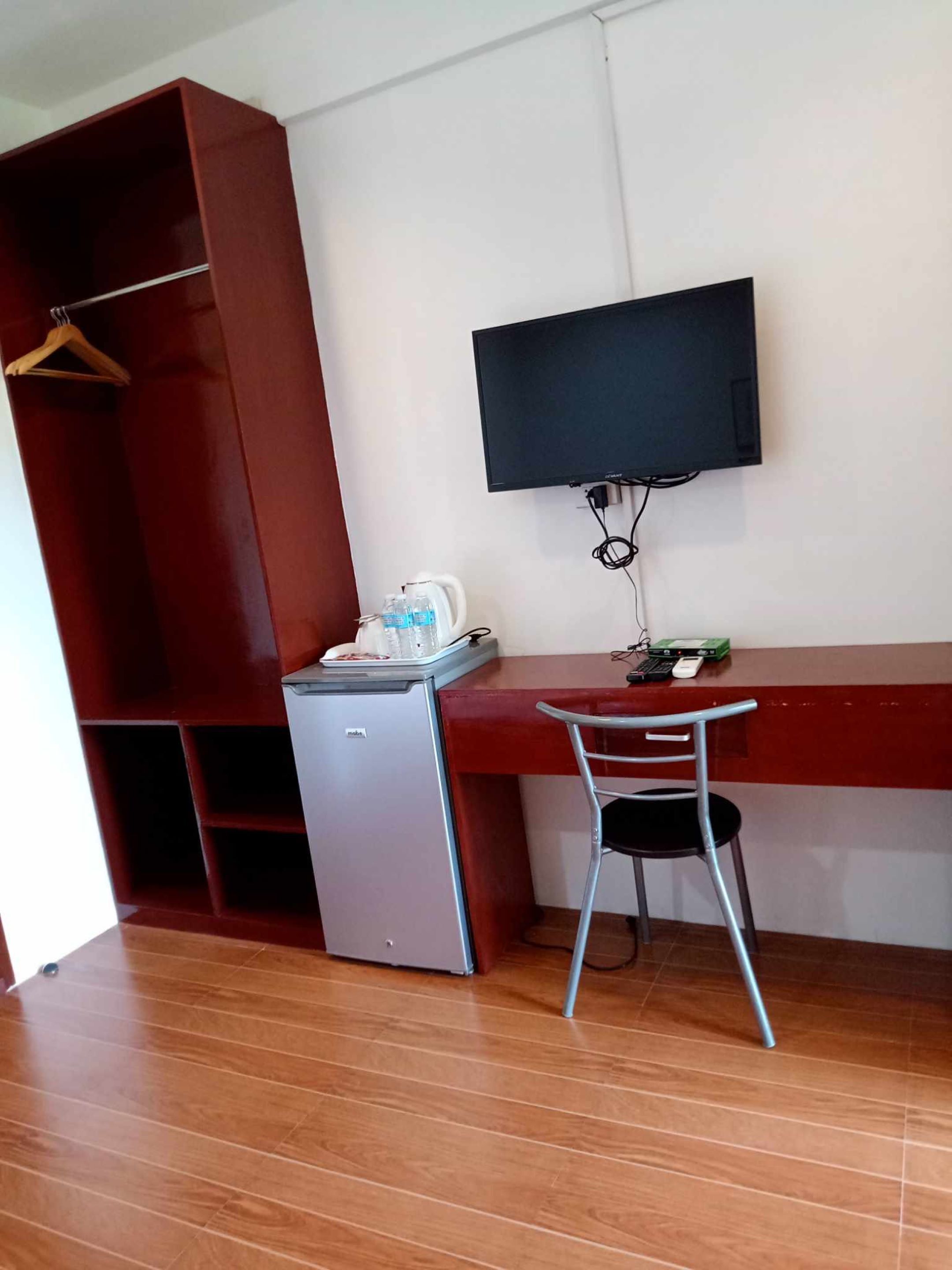 Une pièce de taille modeste, comprenant un bureau en bois, un petit réfrigérateur, une chaise et un téléviseur fixé au mur.