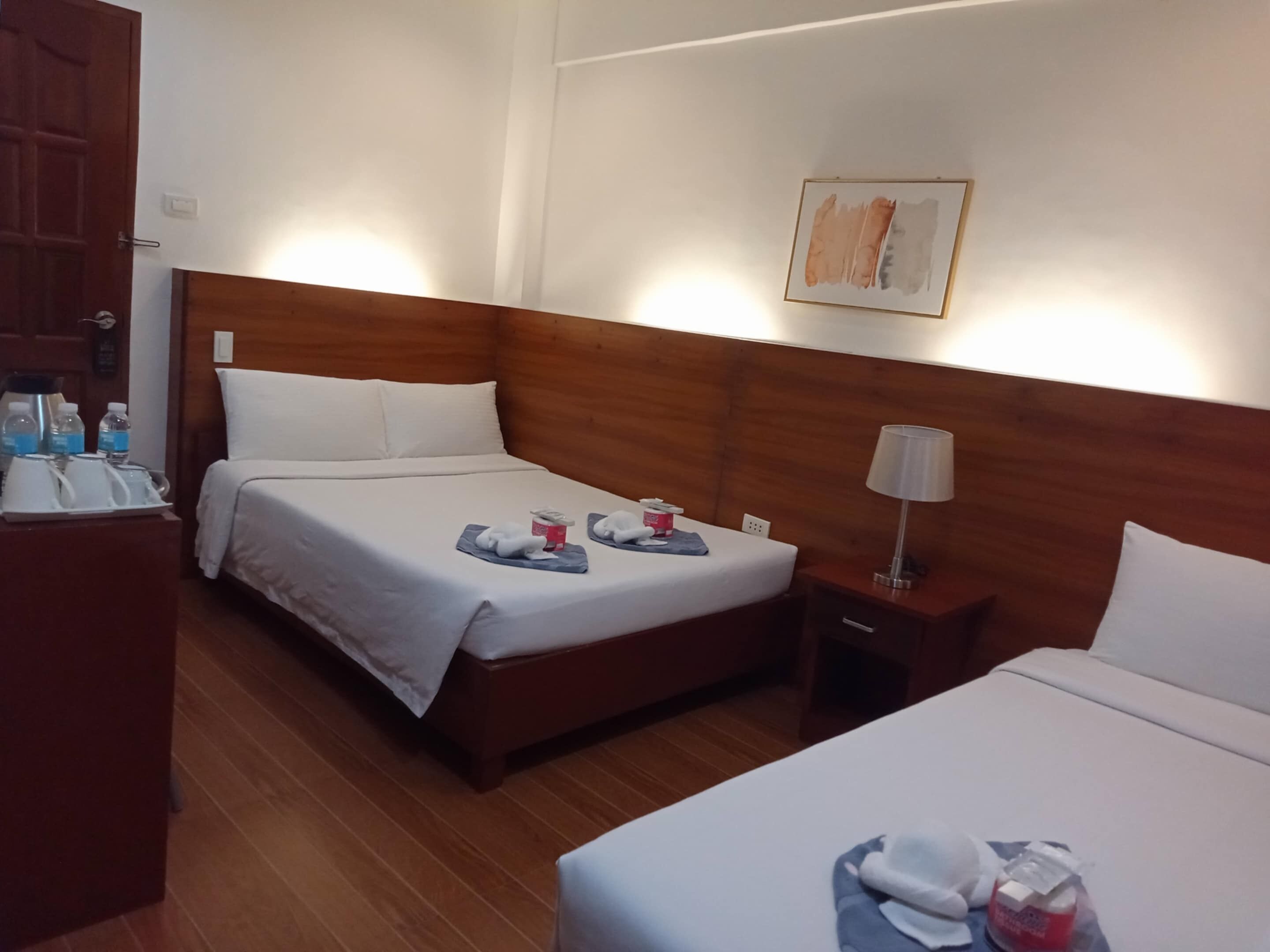 Een hotelkamer met twee bedden, houten hoofdborden, wit beddengoed, een nachtkastje met een lamp en een schilderij aan de muur.