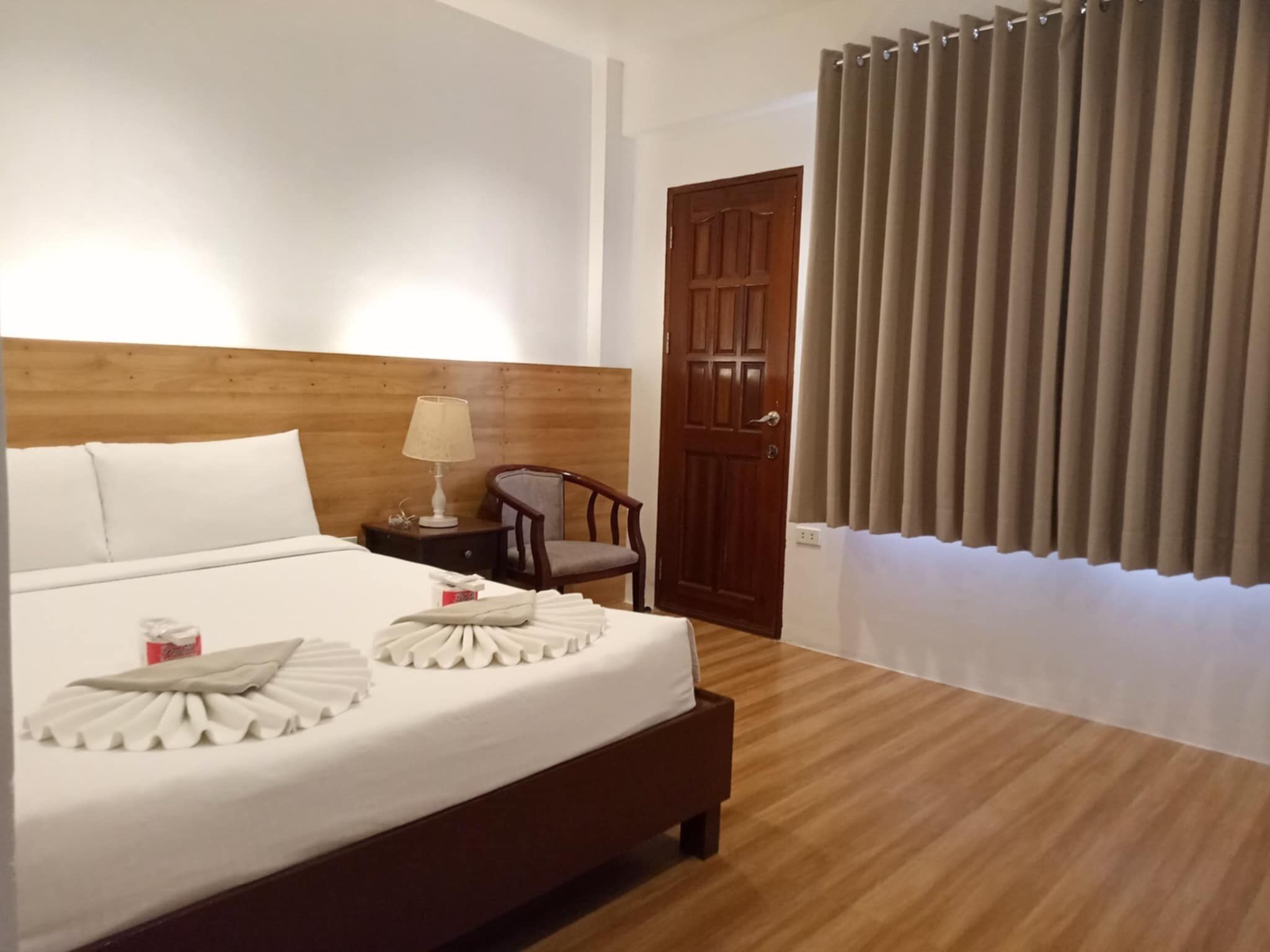 Habitació d'hotel amb un capçal de llit de fusta, un llit amb roba blanca, una meseta de nit amb una làmpada, una cadira i una porta amb cortines.