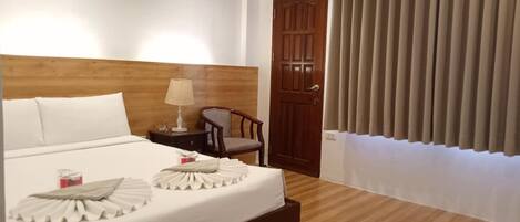 Habitació d'hotel amb un capçal de llit de fusta, un llit amb roba blanca, una meseta de nit amb una làmpada, una cadira i una porta amb cortines.