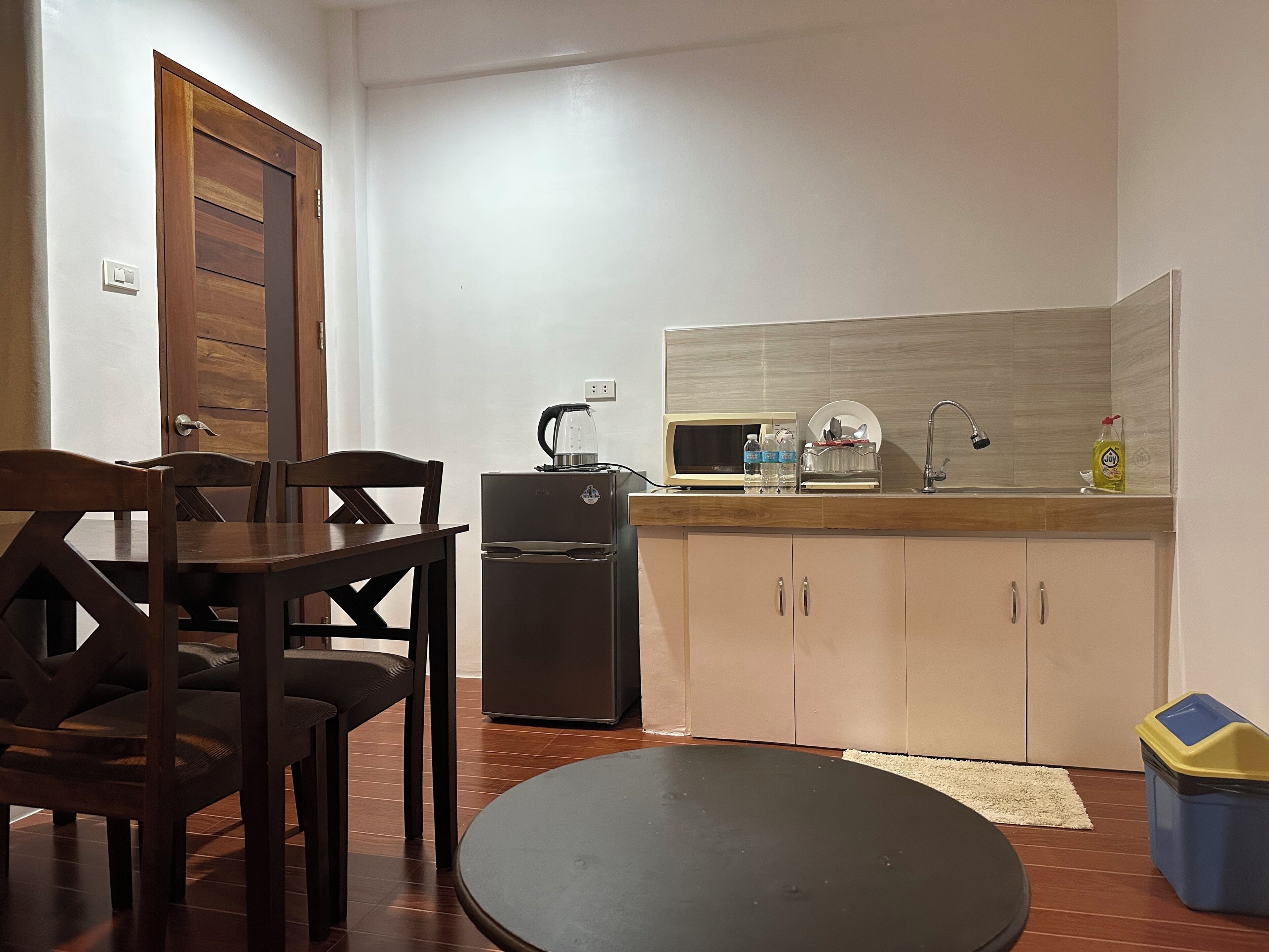 Cocina compacta con refrigerador, microondas y lavamanos. Hay una mesa de comedor con sillas y una pequeña mesa redonda.
