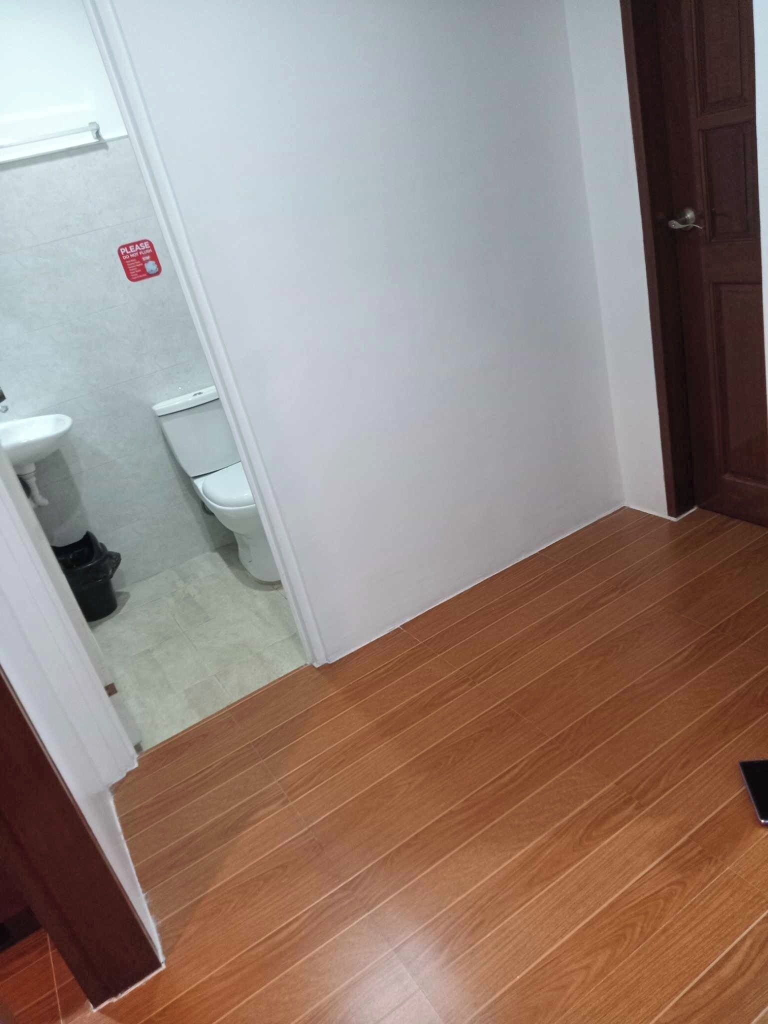Un plancher en bois mène à une porte blanche, avec des toilettes visibles et un panneau rouge en arrière-plan.