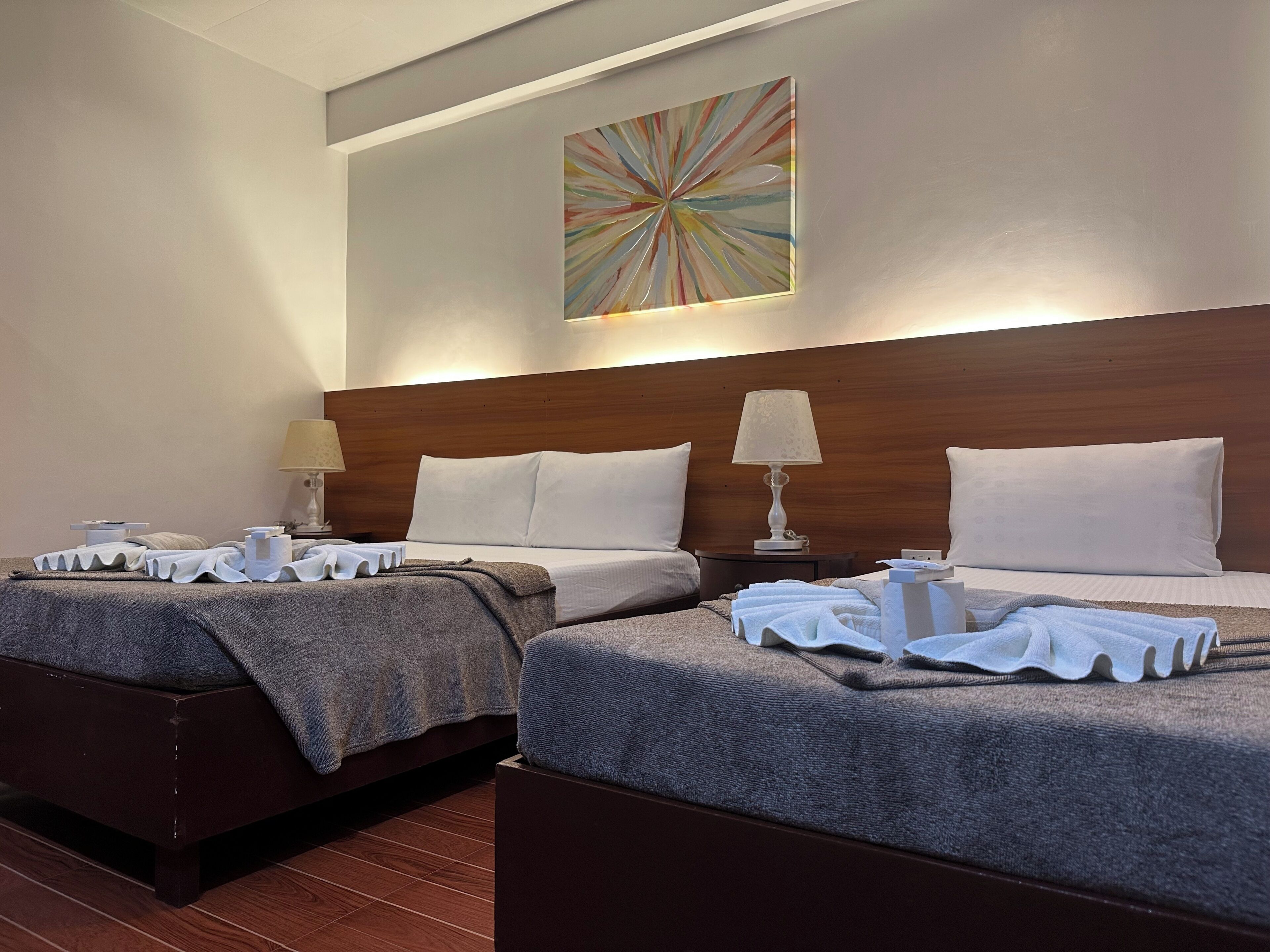 Habitación de hotel con dos camas, cada una con un almohadón blanco y una colcha. Se observa un cabecero de madera, una lámpara fijada a la pared y un cuadro abstracto de colores sobre las camas.