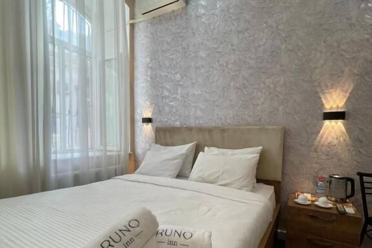 A hotel room with a bed, a nightstand, an air conditioning unit, and a window with curtains.