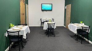 Un salón de conferencias con cuatro mesas, cada una cubierta con manteles blancos y platos con borde dorado. Un televisor de pantalla plana está montado en la pared.