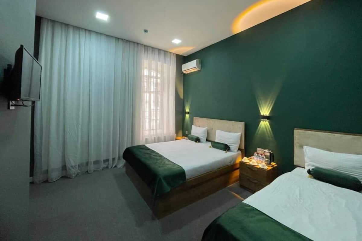 A hotel room with two beds, a flat-screen TV, a window with sheer curtains, and wall-mounted lighting.