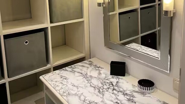 A bathroom with a marble countertop, a large mirror with lights, and a storage unit with open shelves.