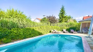 A rectangular swimming pool with a tiled edge, surrounded by a landscaped garden with various plants and a covered seating area with lounge chairs.