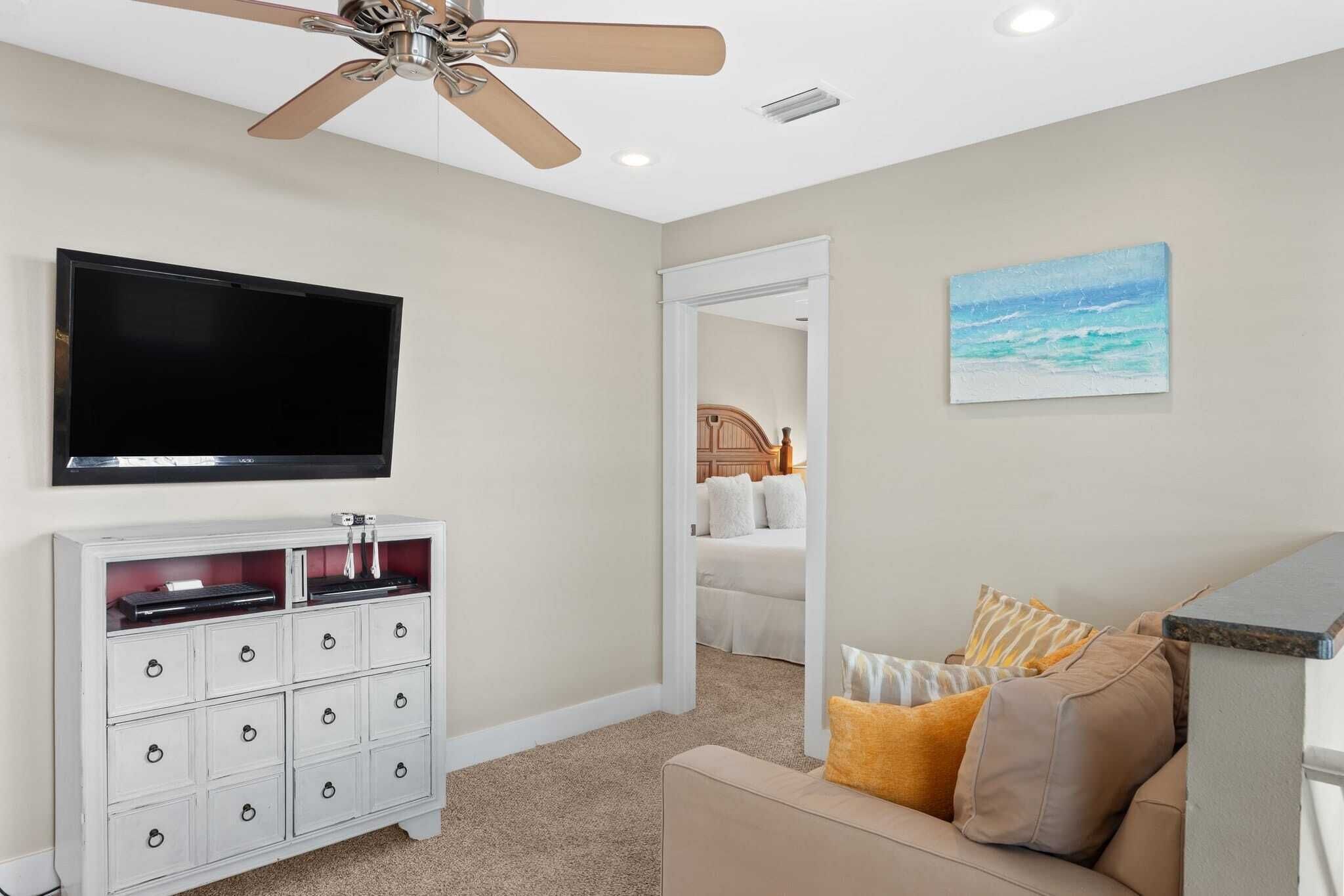 Una stanza con ventilatore a soffitto, televisione a schermo piatto, mobiletto bianco con cassetti, divano beige con cuscini e un quadro raffigurante una spiaggia.