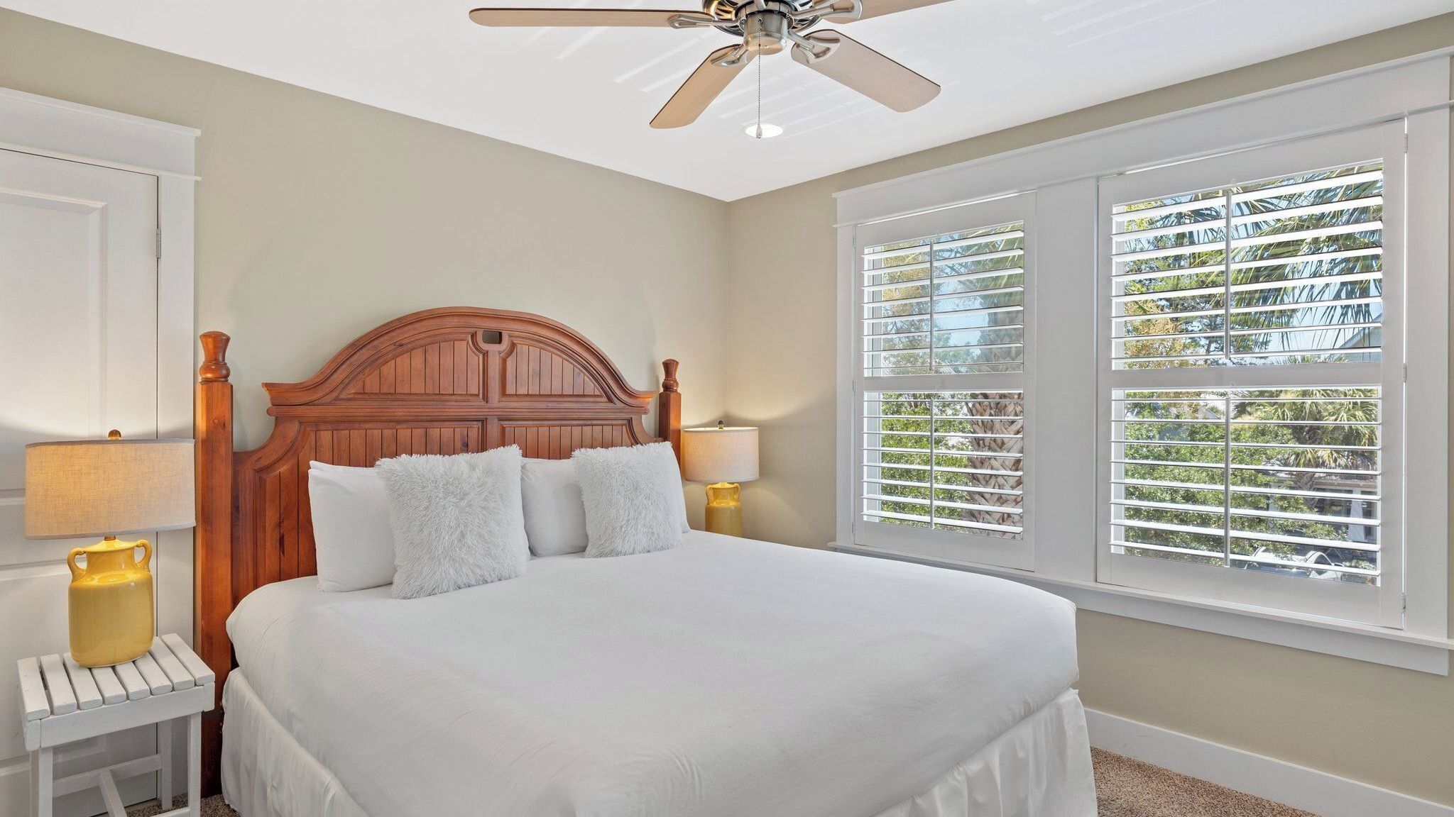A bedroom with a wooden headboard, two bedside lamps, a large window with blinds, and a ceiling fan.