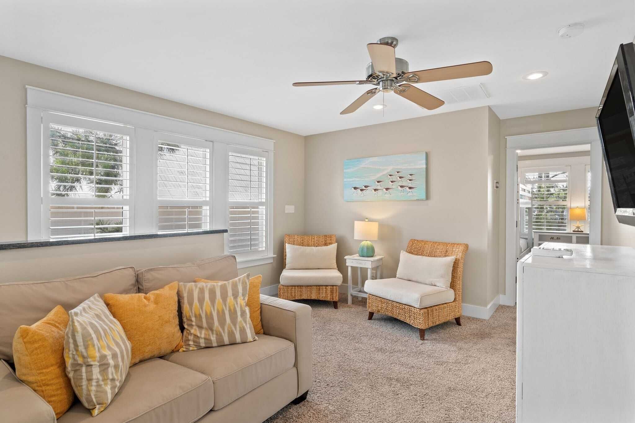 A living room with a beige sofa, two wicker chairs, a ceiling fan, and a painting on the wall.