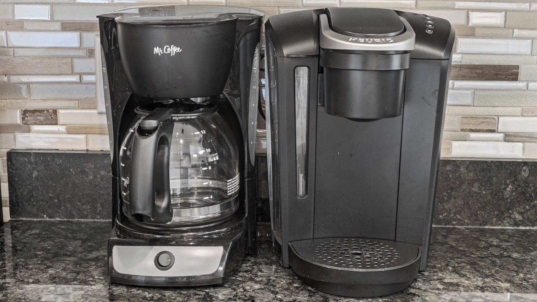 Zwei Kaffeemaschinen, eine mit der Aufschrift 'Mr. Coffee' und die andere 'KEURIG', stehen auf einer Granitarbeitsplatte mit Fliesenrückwand.