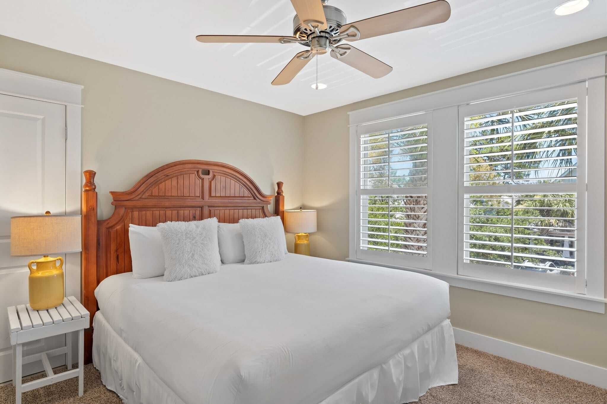 A bedroom with a wooden headboard, white bedding, two bedside lamps, a ceiling fan, and a window with blinds.