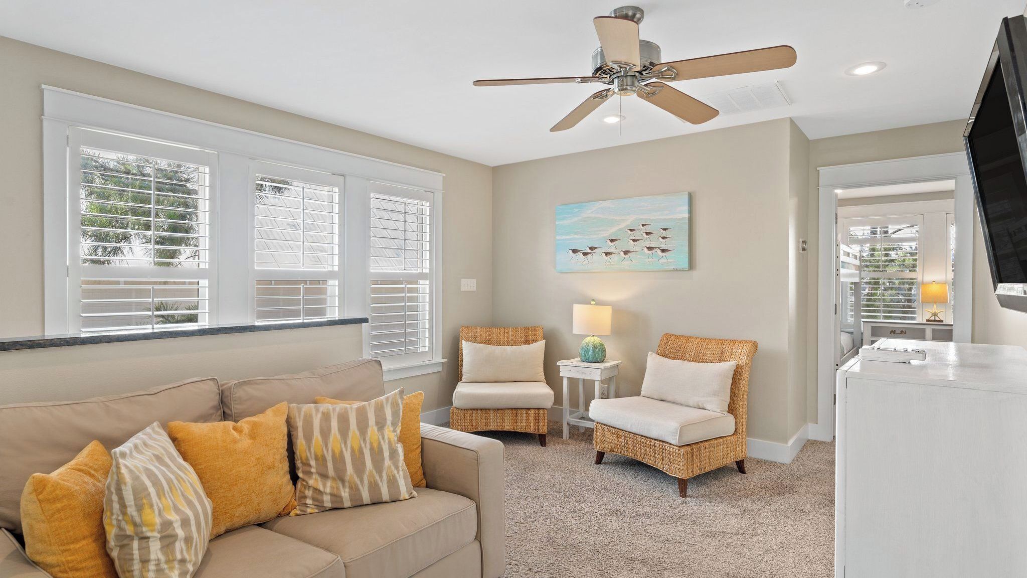 A living room with a beige sofa, two wicker chairs, a ceiling fan, and a painting of birds on the wall.