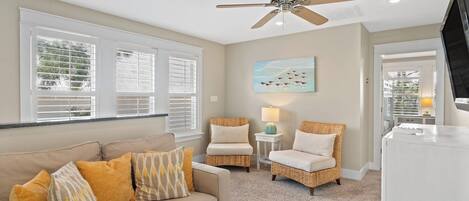A living room with a beige sofa, two wicker chairs, a ceiling fan, and a painting of birds on the wall.
