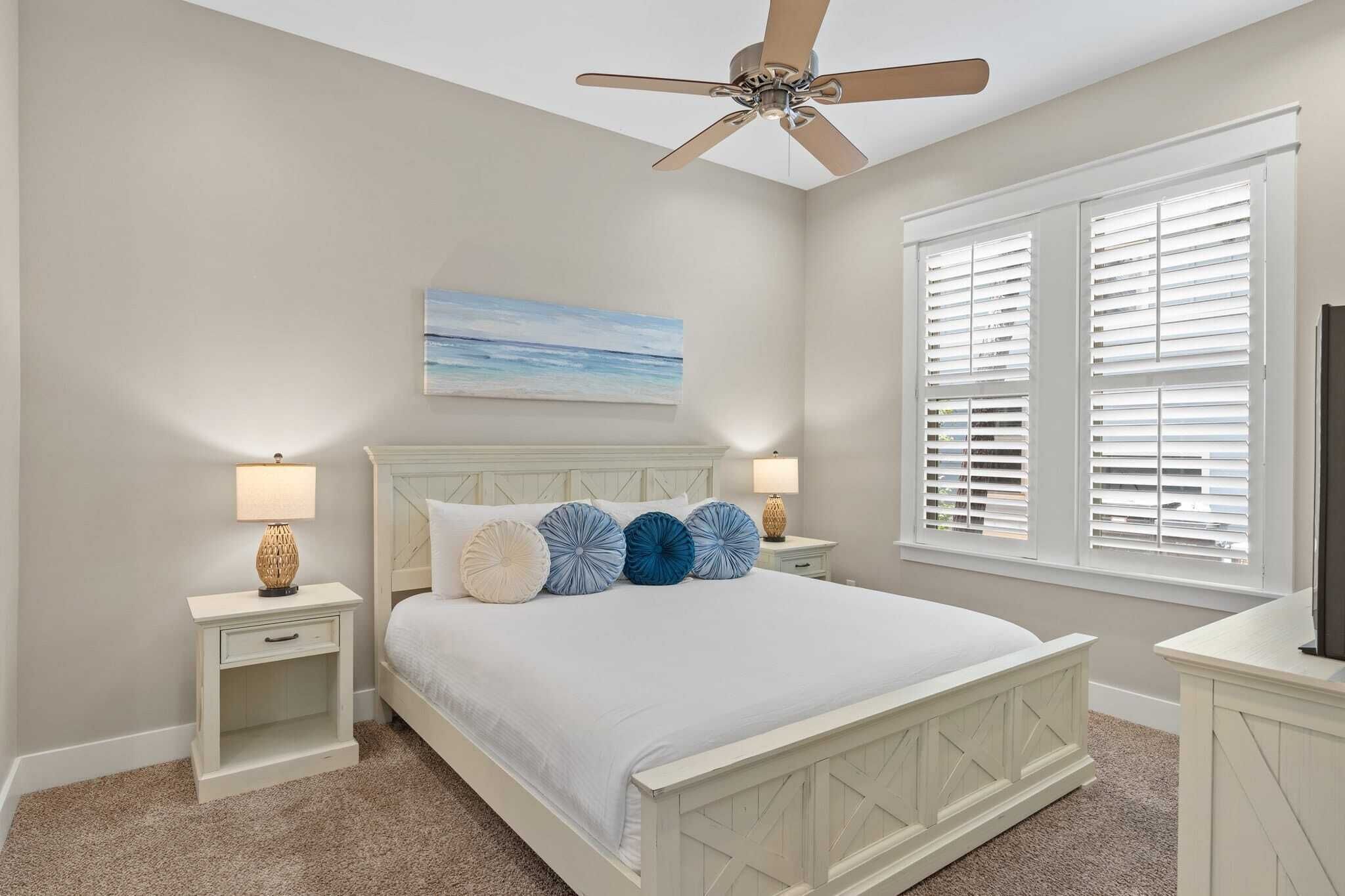 A bedroom with a bed, two bedside tables, a ceiling fan, a window with blinds, and a painting on the wall.