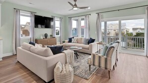 A spacious living room with a beige sofa, a striped armchair, and a coffee table. There is a ceiling fan, large windows with curtains, and a view of the outside. - This is How We Roll- Shared Pool! (surf City)