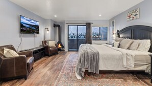 Un dormitorio moderno con una cama grande, una chimenea, un televisor y un balcón con vistas a árboles cubiertos de nieve.