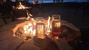 Un hogar con fuego, dos vasos de bebidas, sillas y vista al agua y los árboles.