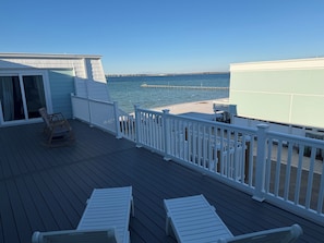 A deck with white railings, a rocking chair, and two lounge chairs overlooking a beach and pier.