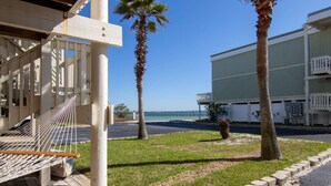 Una hamaca en una terraza de madera, una zona con césped, palmeras y un edificio con balcón con vista al océano.