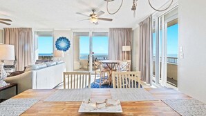 A spacious living area with a wooden dining table, a sofa, and a view of the ocean. - 4 BR Top Floor Beachfront Condo | Beach Club B2010 (Baldwin County)