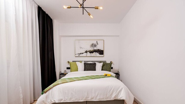 A bedroom with a bed, a bedside table, a lamp, and a wall-mounted artwork. - Stylish City Living Modern Apartment in the hear of Bucharest (București)