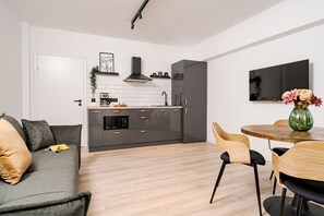 A modern kitchen with a built-in oven, microwave, and sink, a dining area with a round table and chairs, and a grey sofa with cushions. - Tyst by MRG Apartments | City Centre Spacious Cozy Apartment with Balcony (București)