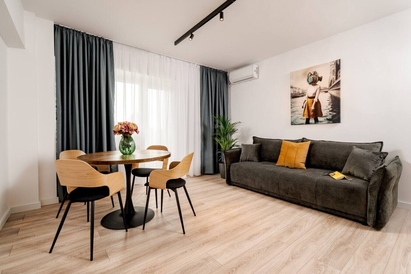 A modern living room with a dining area, a grey sofa, a painting on the wall, and a potted plant.