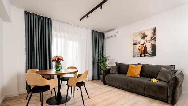 A modern living room with a dining area, a grey sofa, a painting on the wall, and a potted plant. - Tyst by MRG Apartments | City Centre Spacious Cozy Apartment with Balcony (București)