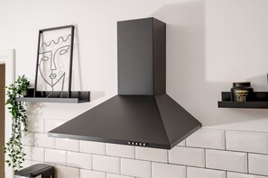 A kitchen with a modern range hood, white tiles, and a minimalist decor including a framed artwork and a small shelf with a jar.