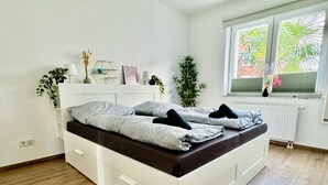 Moderni makuuhuone, jossa on valkoinen sänky, puulattia ja ikkuna, josta avautuu näkymä vehreään maisemaan.