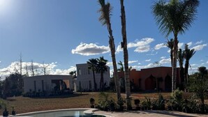 A clear blue pool with a curved edge, surrounded by palm trees and a building with a flat roof in the background. - Yin Hotel (Marrakech)
