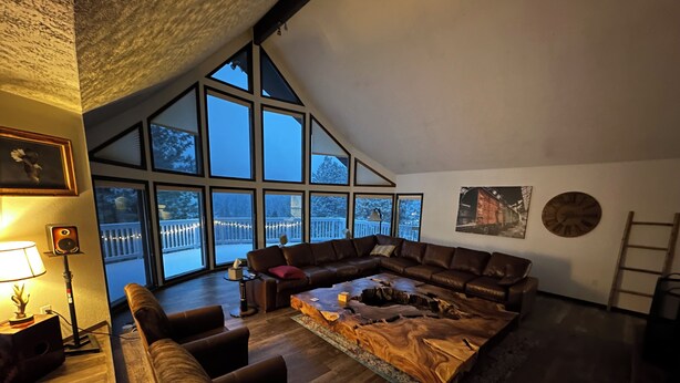 Un salón espacioso con un sofá modular en forma de L, una mesa de centro de madera y una chimenea. Amplias ventanas ofrecen vistas a un balcón con guirnaldas luminosas.