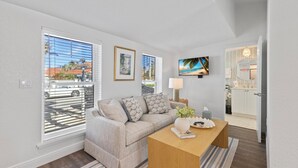 A modern living room with a sofa, a coffee table, and a TV. There is a window with blinds, a framed picture, and a door leading to a bathroom. - Anchor Inn 02 | 1BR/1BA Boca Grande, Chic Comfort (Boca Grande)
