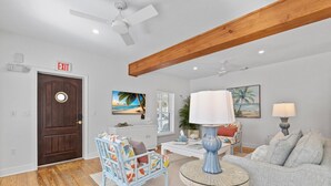 A spacious living room with a wooden ceiling, a large door marked 'EXIT', a blue armchair, a grey sofa, and a round table with a lamp.