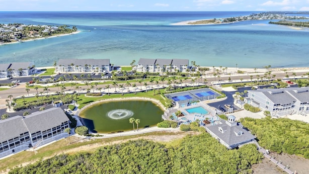 An aerial view of a coastal resort with multiple buildings, a central pool, and a large body of water.