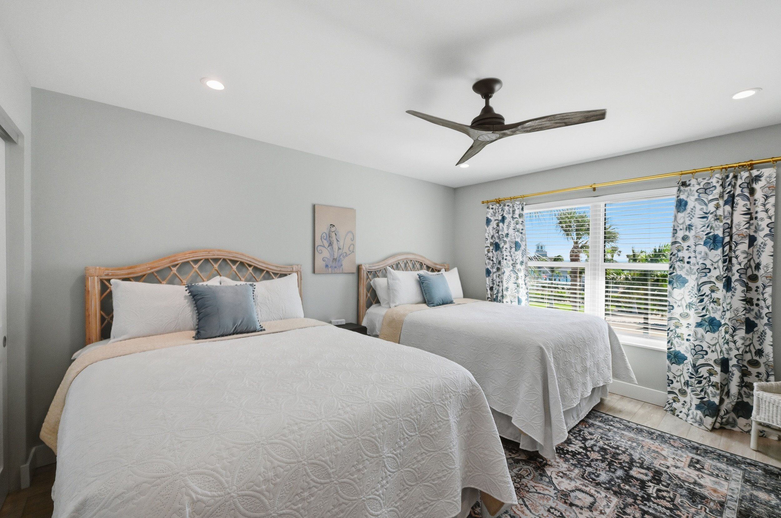 A bedroom with two beds, a ceiling fan, a window with curtains, and a painting on the wall.