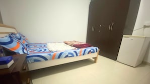 A single bed with a colorful bedspread, a small bedside table with a book, a wardrobe with two doors, and a white refrigerator.