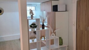 A room with a white shelving unit, a wooden wardrobe, and a small white cabinet with a microwave on top.