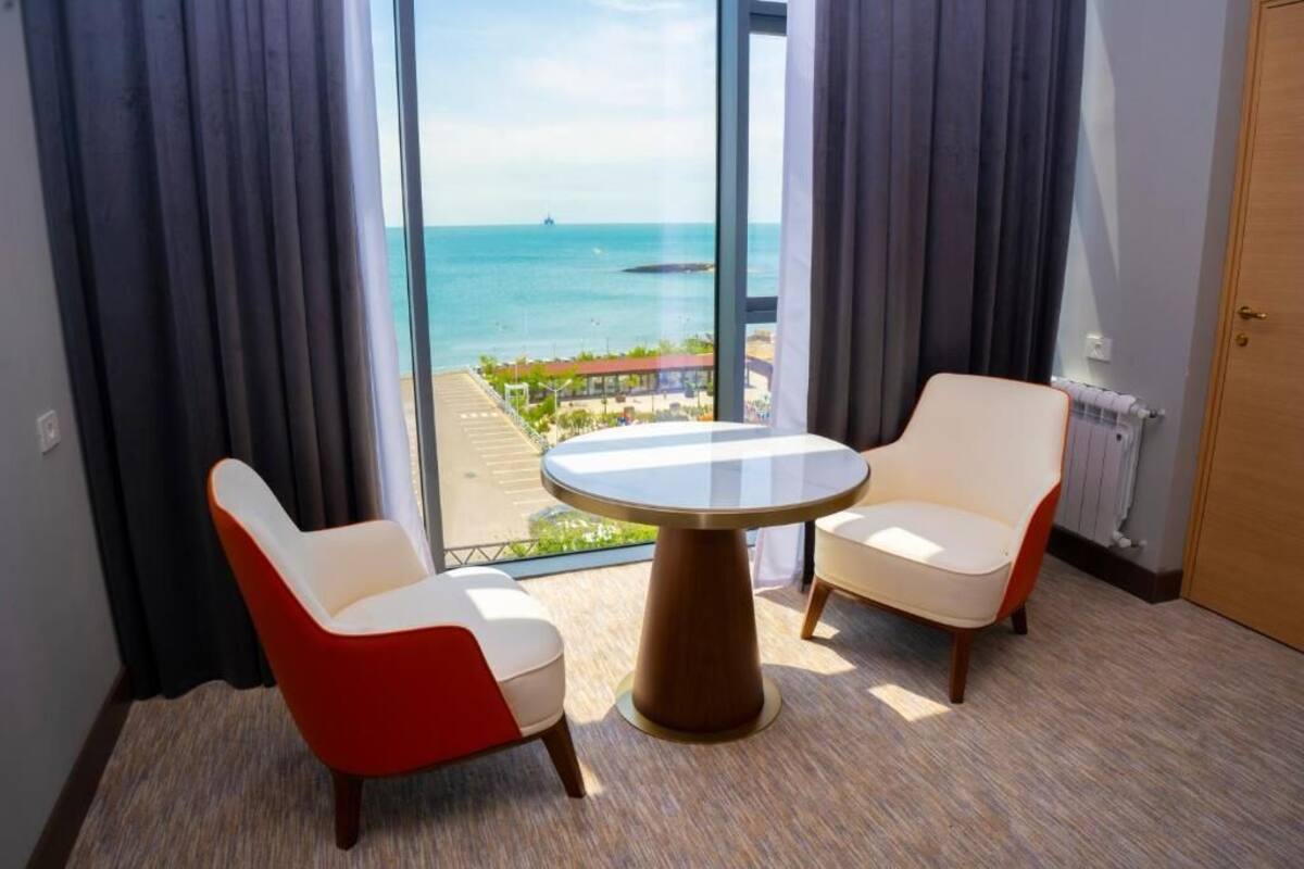 A room with a large window offering a view of the sea and a distant cityscape. There is a round table with two chairs, one red and one white, in the center of the room.