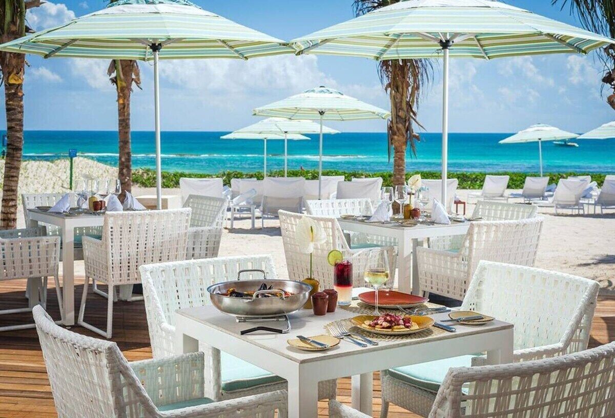 A beachside dining area with white wicker furniture, umbrellas, and a view of the ocean.