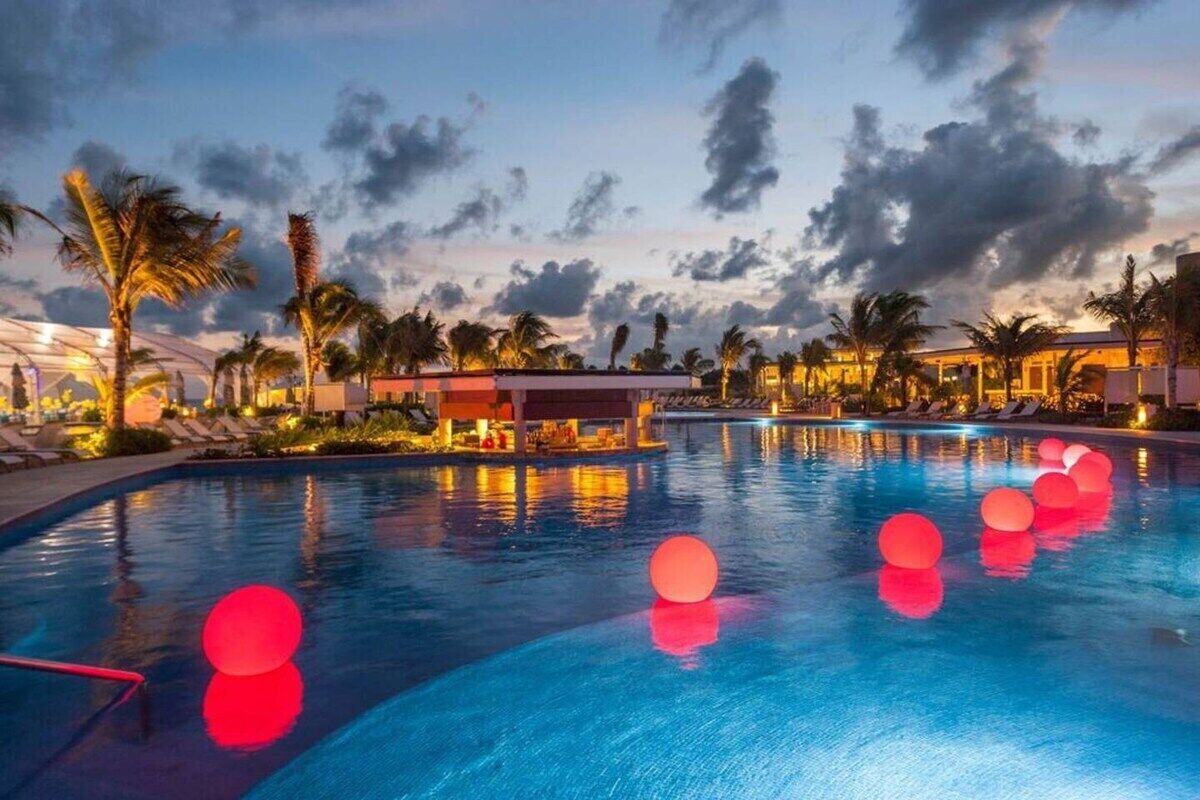 Una piscina con pelotas rojas flotando, palmeras y un edificio con techo rojo al fondo.