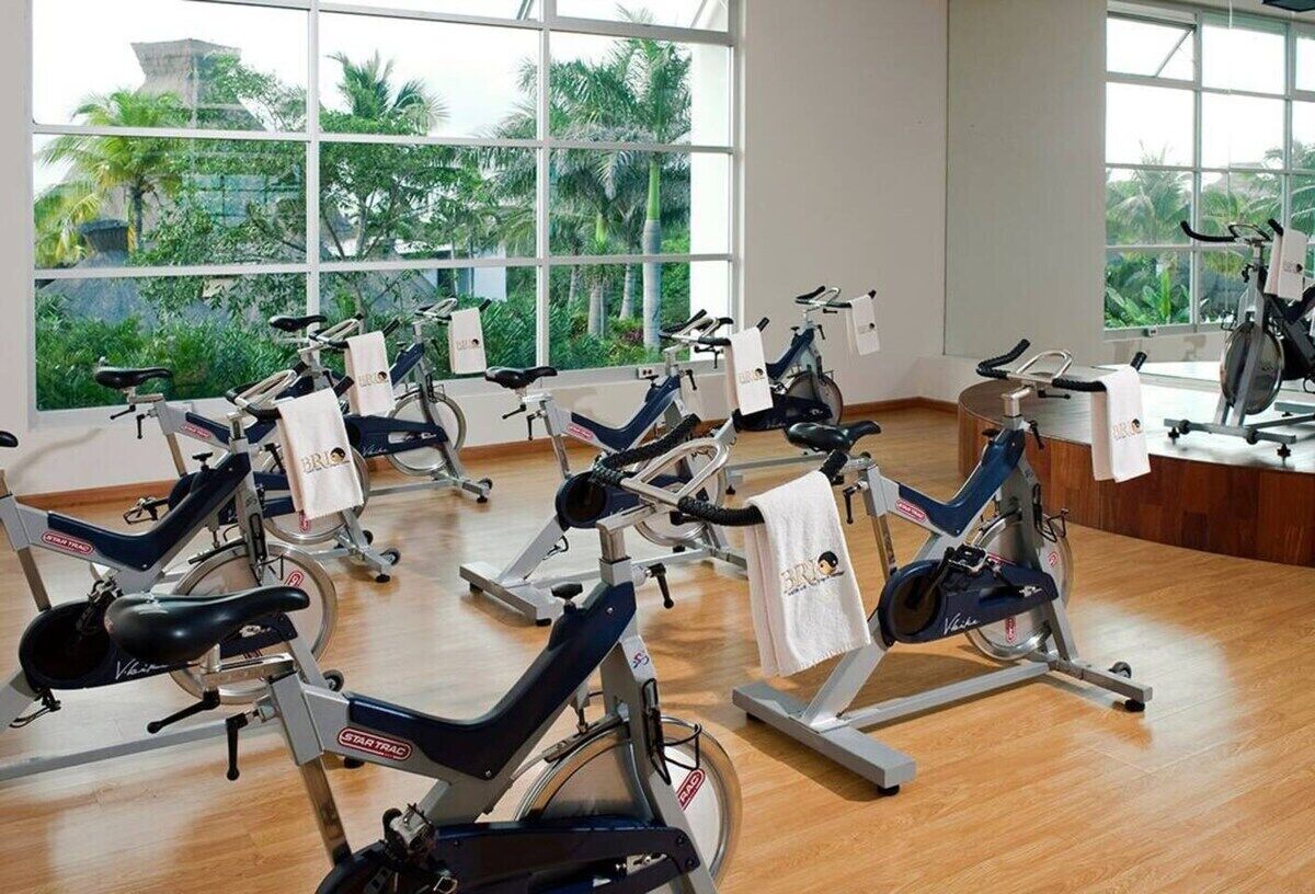 A gym with stationary bikes arranged in rows, large windows with a view of palm trees, and a wooden floor.