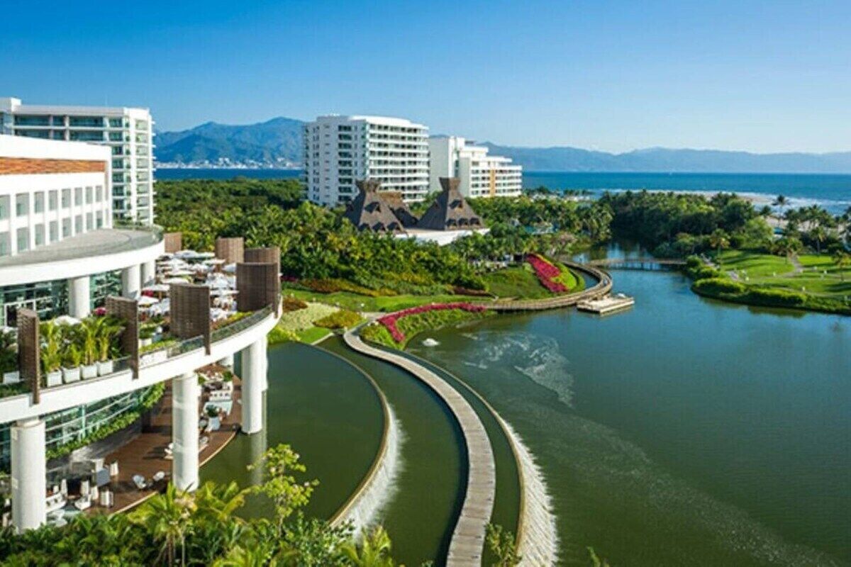 Un hotel moderno con un canal curvo, abundante vegetación y una vista de montañas al fondo.