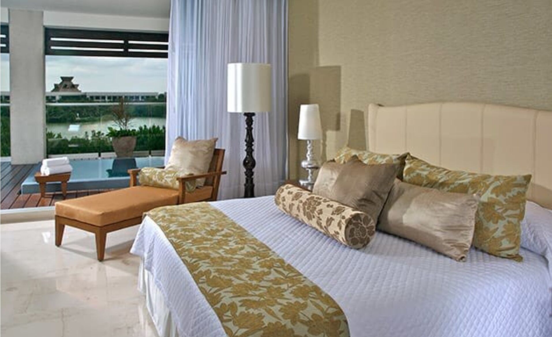 A hotel room with a bed, a chair, a table, and a view of a building and greenery.