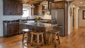 A kitchen with wooden cabinets, a stainless steel refrigerator, and a central island with stools. - Gull Lake Cabin + Bunkhouse with Sandy Beach & Pontoon/Jet Ski availability! (Nisswa)