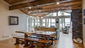 A spacious living area with a wooden dining table, benches, and a sofa. There are large windows providing natural light and a view of the outdoors. - Gull Lake Cabin + Bunkhouse with Sandy Beach & Pontoon/Jet Ski availability! (Nisswa)