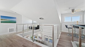A spacious loft apartment with a high ceiling, a balcony with glass railings, a dining area with a bar, and a large TV mounted on the wall.