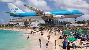 A large blue and white airplane with "KLM asia" written on its tail, flying over a crowded beach with people and umbrellas.